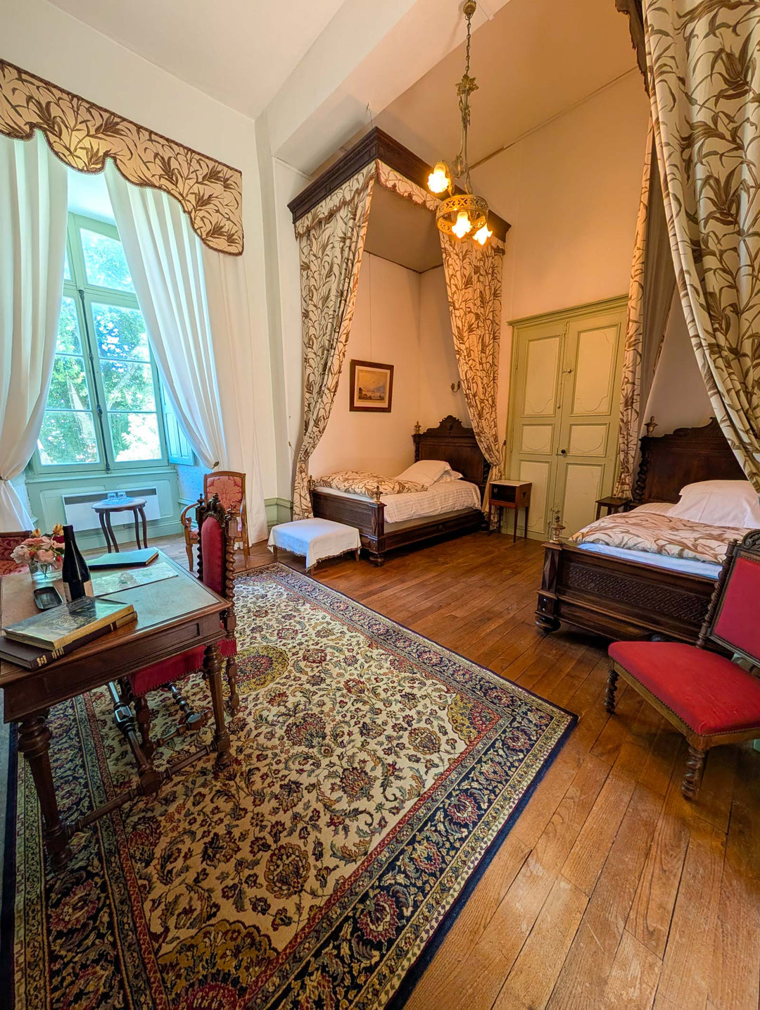 A vintage-style bedroom interior with two wooden beds, patterned canopies, a large ornate rug, a desk with red upholstered chairs, hardwood floors, and a window with sheer curtains.