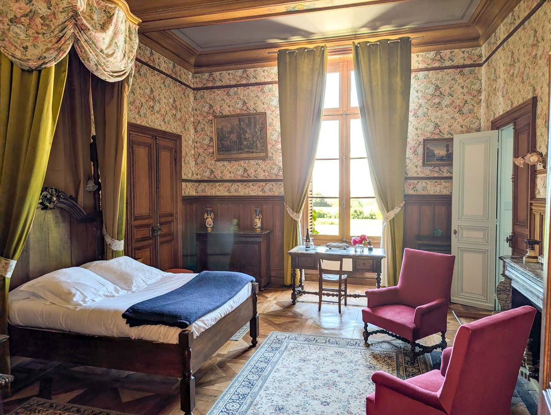 An ornate vintage bedroom with a four-poster canopy bed, floral wallpaper, green drapes, a blue throw blanket, two pink armchairs, and a large patterned rug.