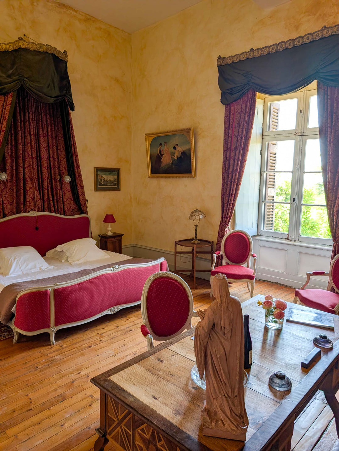 A vintage-style bedroom with a red upholstered bed, matching red and pink chairs, dark red curtains, a wooden table in the foreground with a statue, and warm yellow walls.