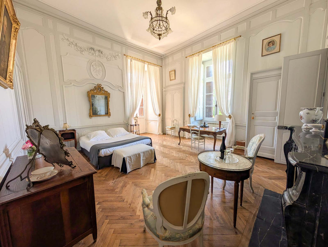 A grand, elegant bedroom interior with a double bed, wooden herringbone floor, ornate white walls, sheer curtains, a chandelier, and multiple antique-style chairs and tables.