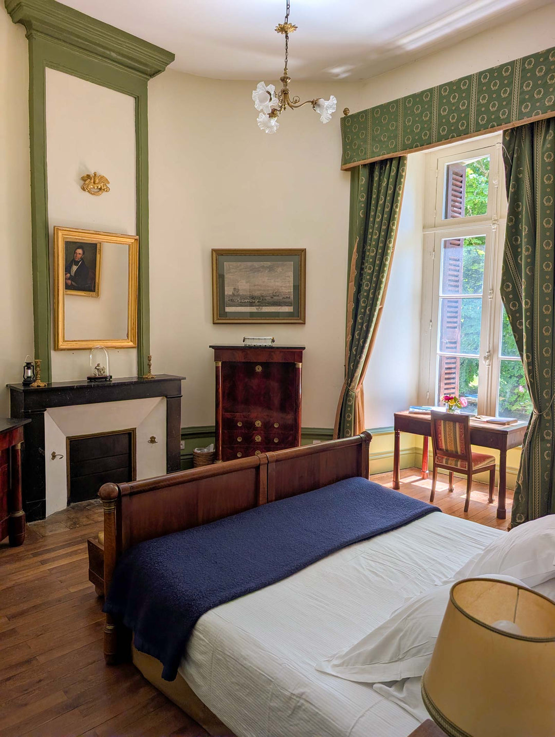 A classic vintage bedroom featuring a wooden bed with white and navy bedding, a dark wood chest, a fireplace with a gold mirror, and green patterned curtains by a large window.