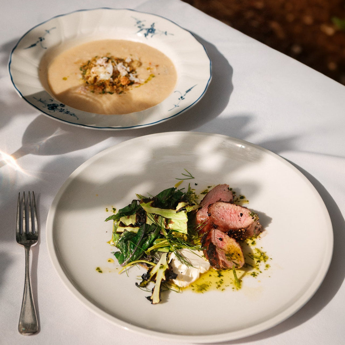 A plated gourmet dish of sliced pink meat with greens and mushrooms on a white plate, alongside a bowl of creamy soup with a fork on a white tablecloth.
