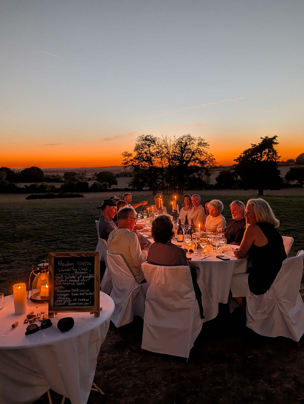 An outdoor evening wedding reception scene with a group of guests seated around a dining table lit by candles, set against a sunset sky over a grassy field.