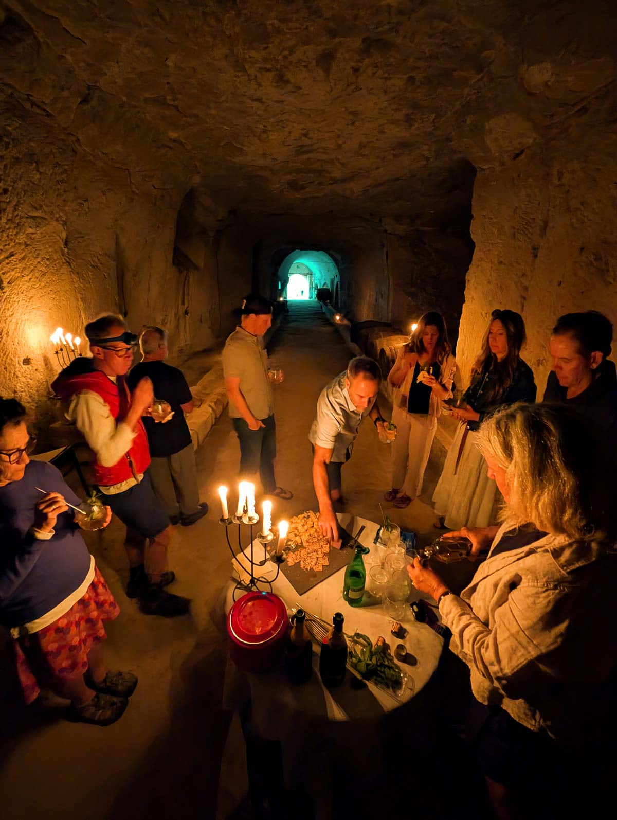 A group of people gathered in a dimly lit stone tunnel, holding candles and drinks around a small table in a rustic, cave-like setting.