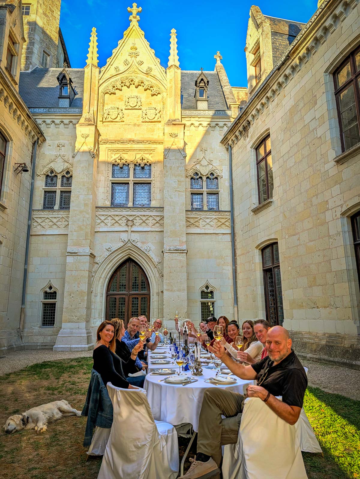 A group of people celebrating with drinks and toast at an outdoor dining table set in a stone chateau courtyard in front of a grand Gothic-style building, with a white dog lying on the grass nearby.