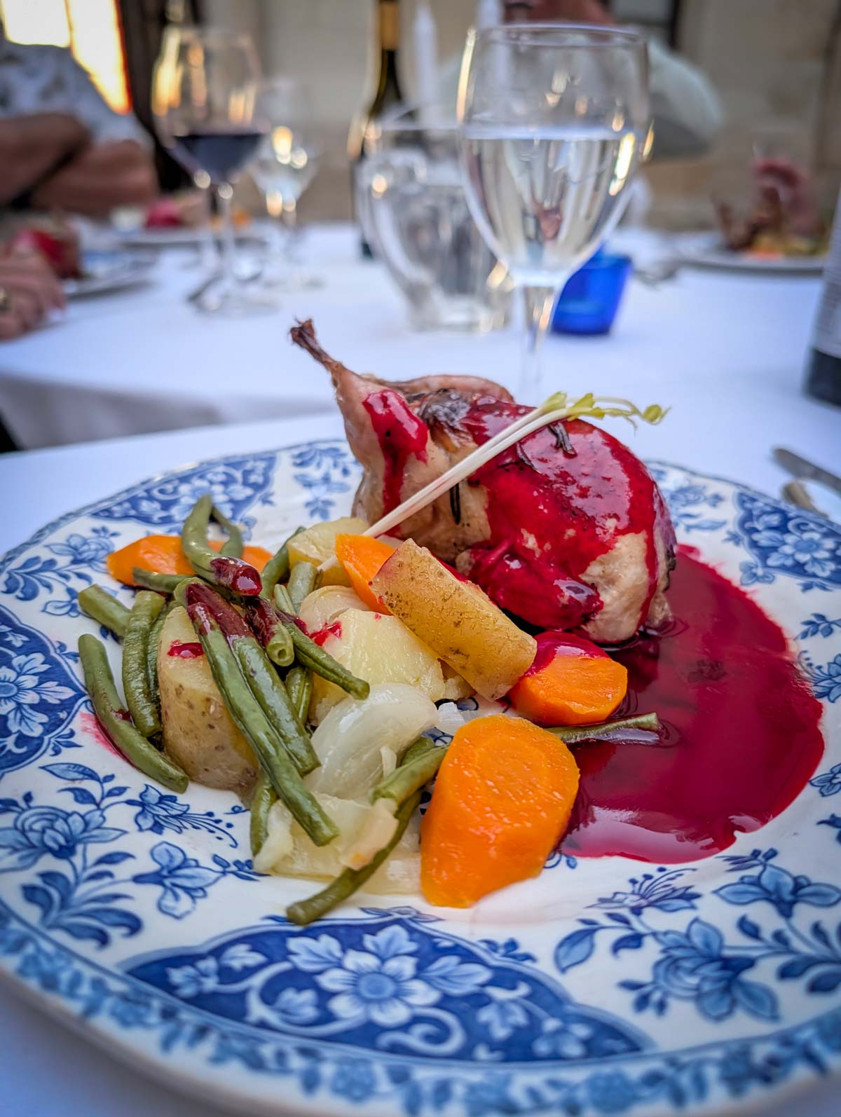 A plated gourmet dish featuring a roasted bird with a dark red sauce, served with green beans, potatoes, and carrots on a blue and white patterned ceramic plate, set on a dining table with wine glasses in the background.