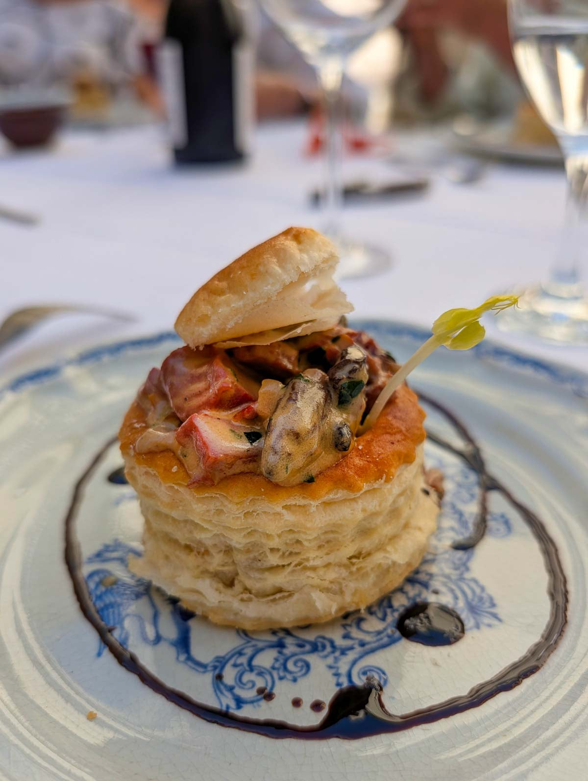 A plated gourmet food dish featuring a flaky pastry stack topped with mushrooms, red fruit, and sauce, set on a blue and white ceramic plate at a dining table.