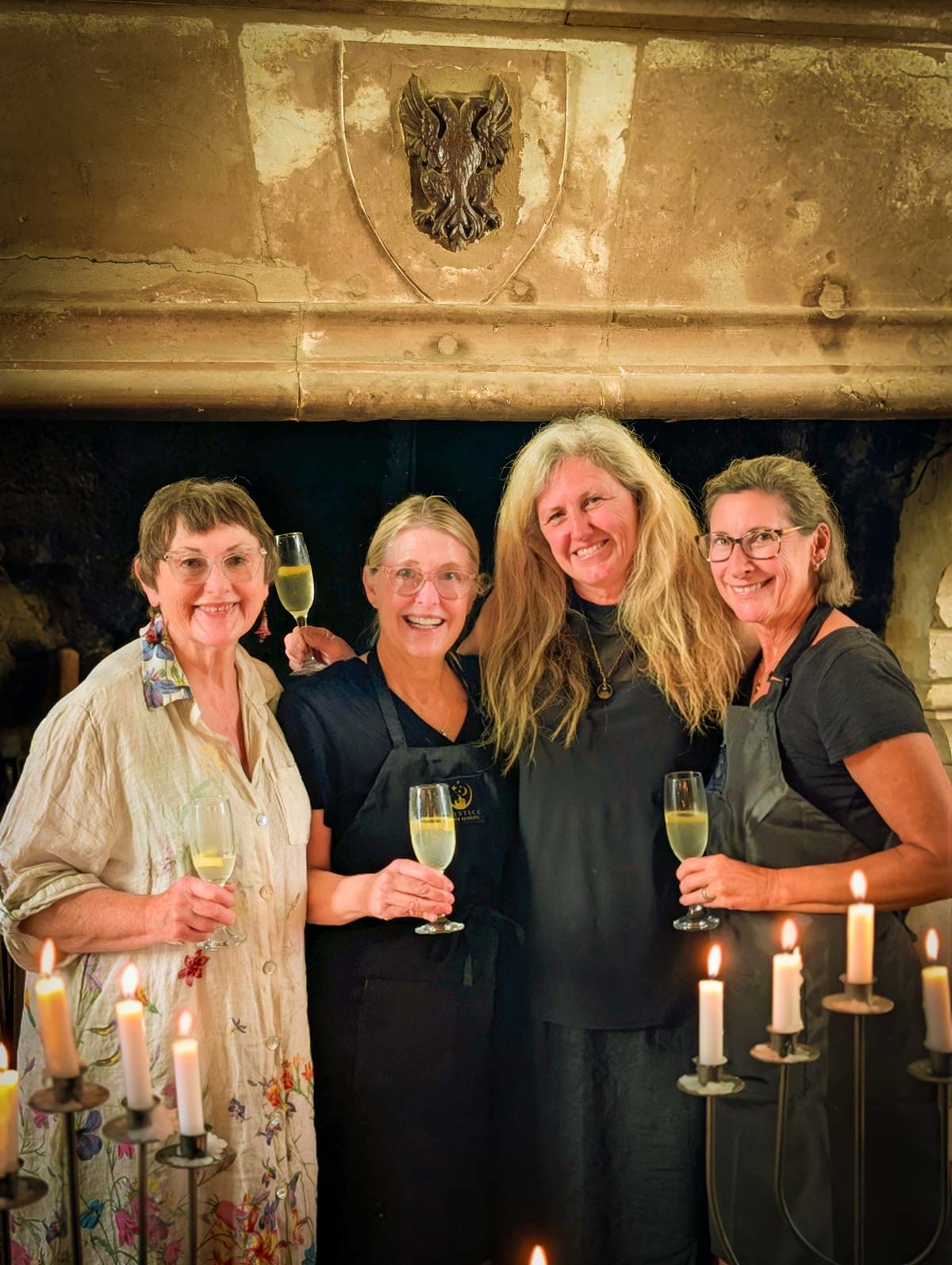Four women standing together in a rustic stone room holding wine glasses, with candles in the foreground and a shield emblem on the wall behind them.