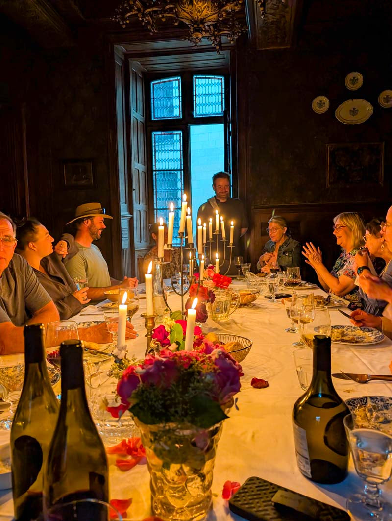 A group of people gathered around a long dining table lit by numerous candles in a dark, elegant room. The table is set with wine bottles, glasses, plates, and a floral centerpiece with pink and purple flowers.