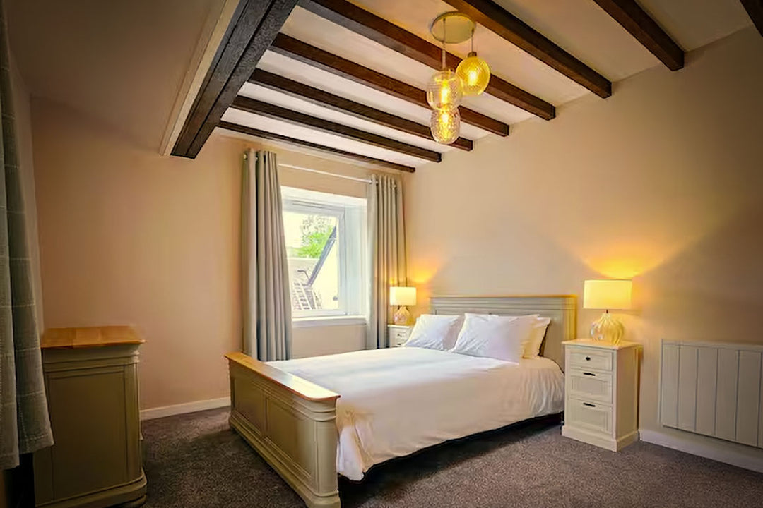 A modern bedroom interior with a bed dressed in gray bedding and a dark blue pillow, a blue upholstered armchair, a wooden nightstand with a lit lamp, and a window with a gray blind. The room has light gray walls, white trim, and a gray carpet.