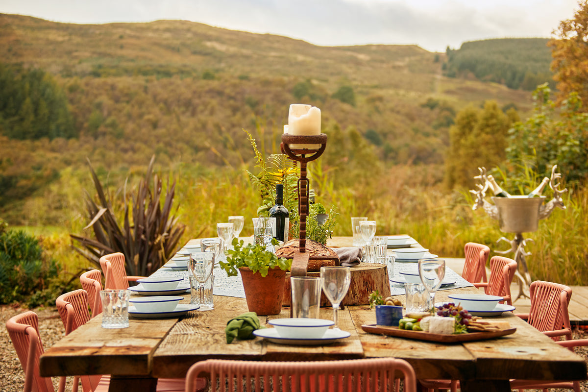 A Scottish rustic outdoor dining table set with blue and white plates, wine glasses, a candle holder, and greenery, surrounded by pink chairs against a backdrop of rolling hills.