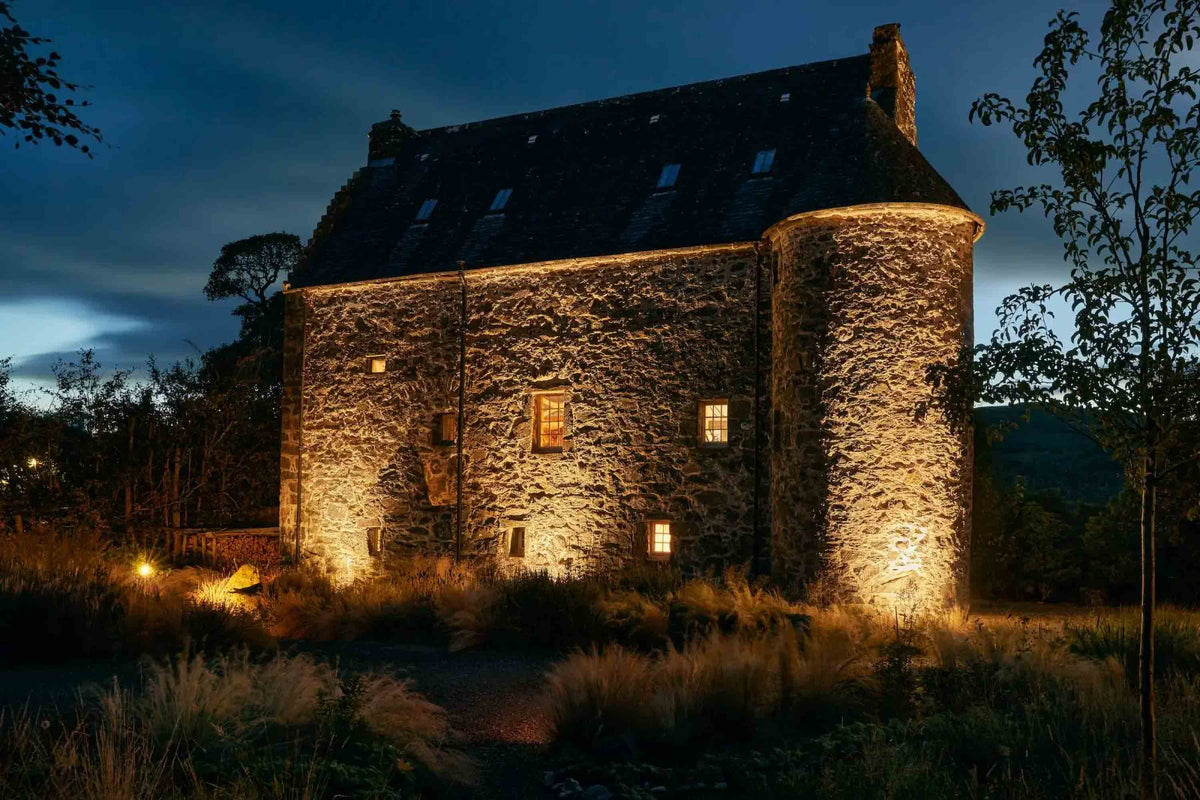 A Scottish stone castle at dusk, warmly lit from within and by exterior ground lights, surrounded by dark trees and grass under a deep blue evening sky.