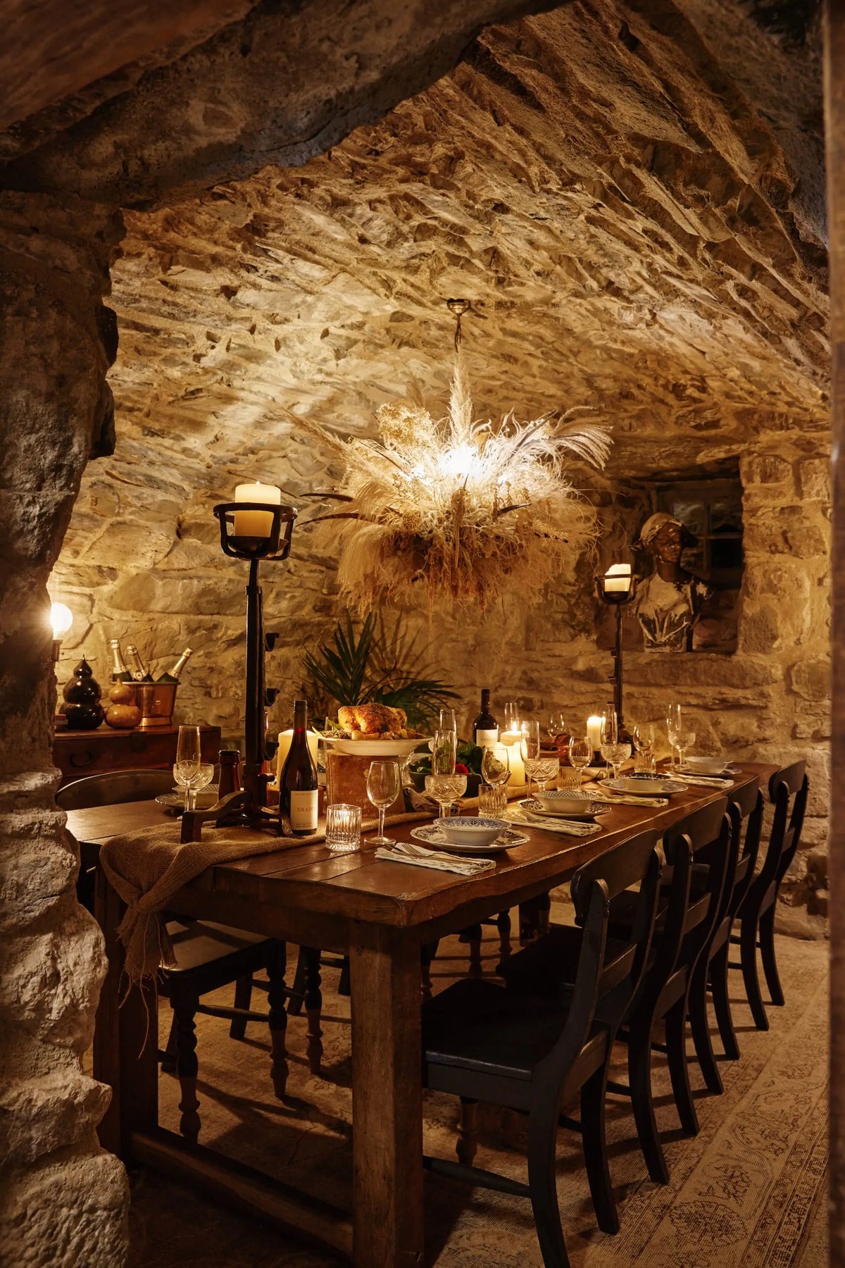 A rustic stone dining room with a long wooden table set for guests, black chairs, candlelit black lanterns, and a warm hanging light fixture against a textured stone wall.