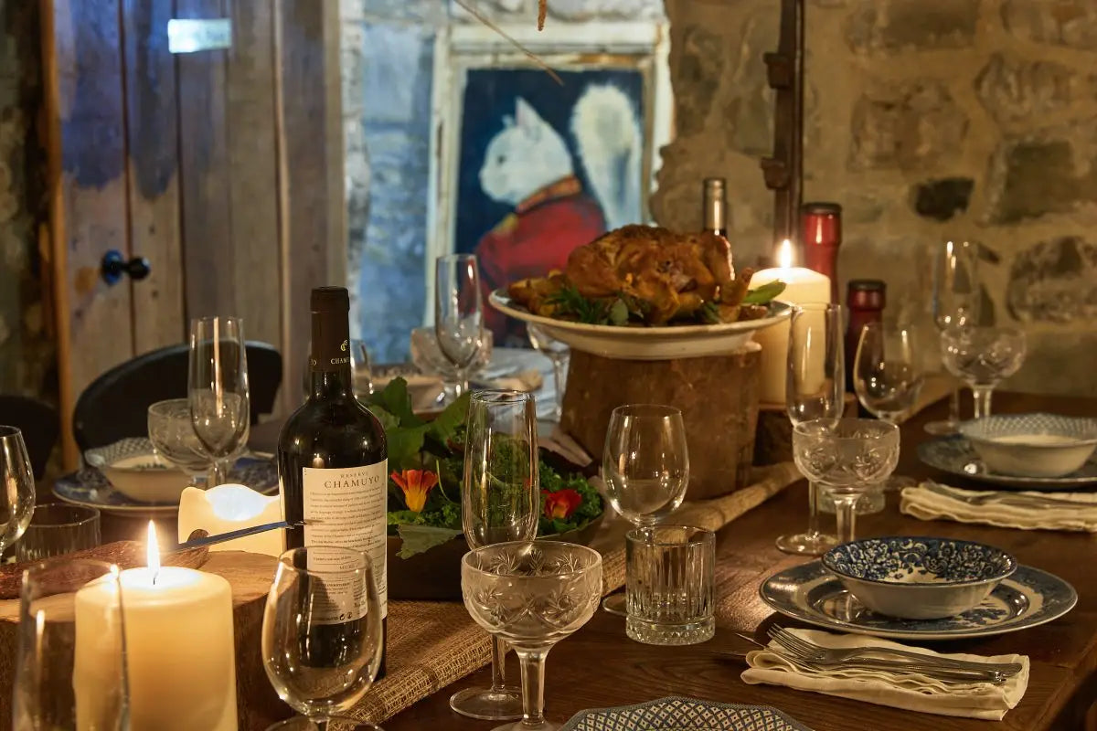 A rustic dining table set for a meal in a stone-walled room, with candles, wine bottles, glassware, plates, and a roasted bird on a platter in the background.