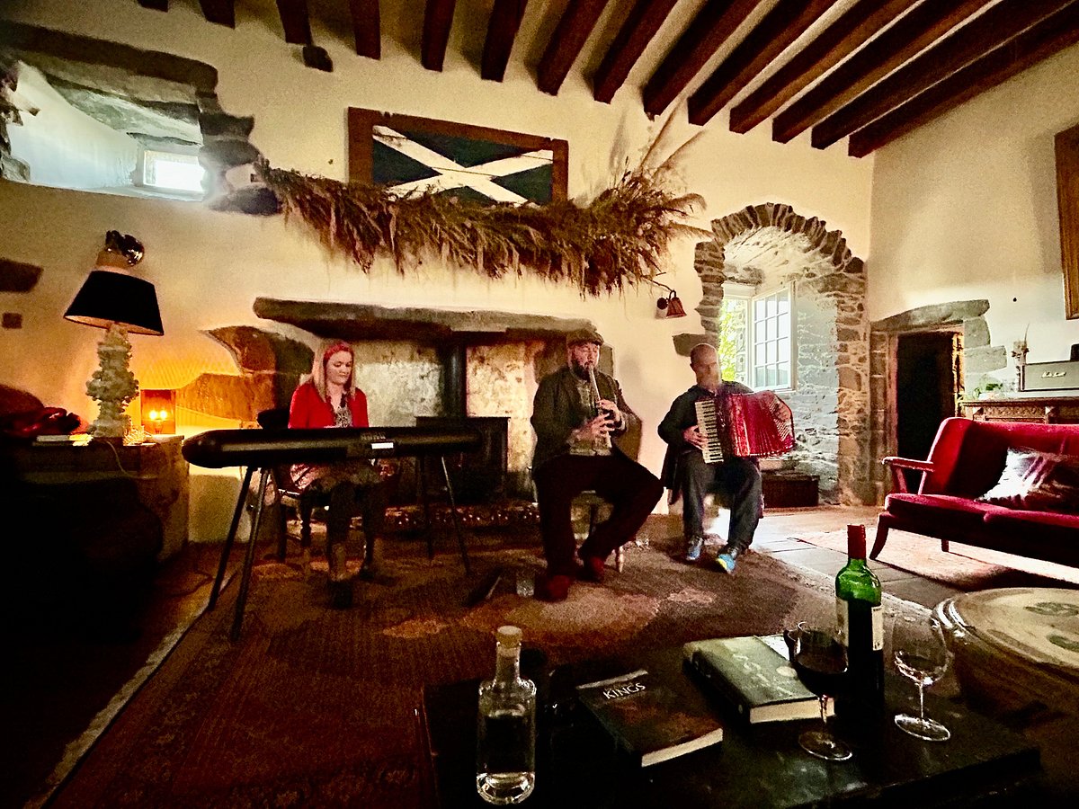 Three musicians playing traditional instruments in a rustic, warmly lit stone room with wooden ceiling beams, a fireplace, and a red sofa in the background.