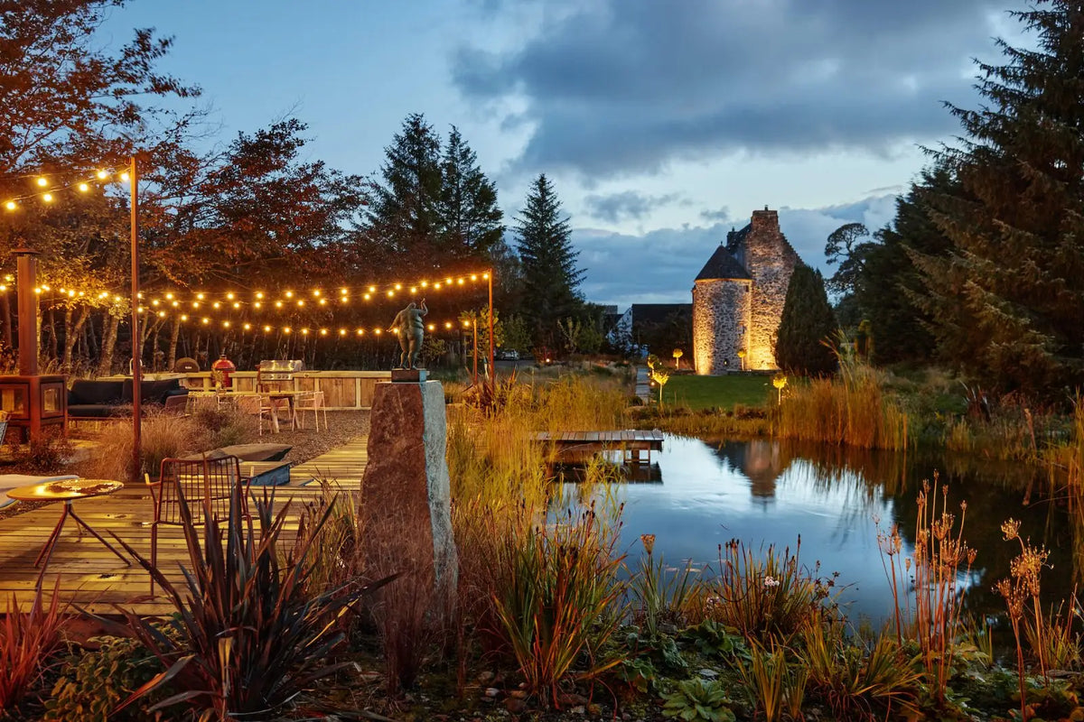 An outdoor evening scene at a rustic stone castle beside a small pond, with warm string lights over a patio area, trees, reeds, and a stone monument in the foreground.