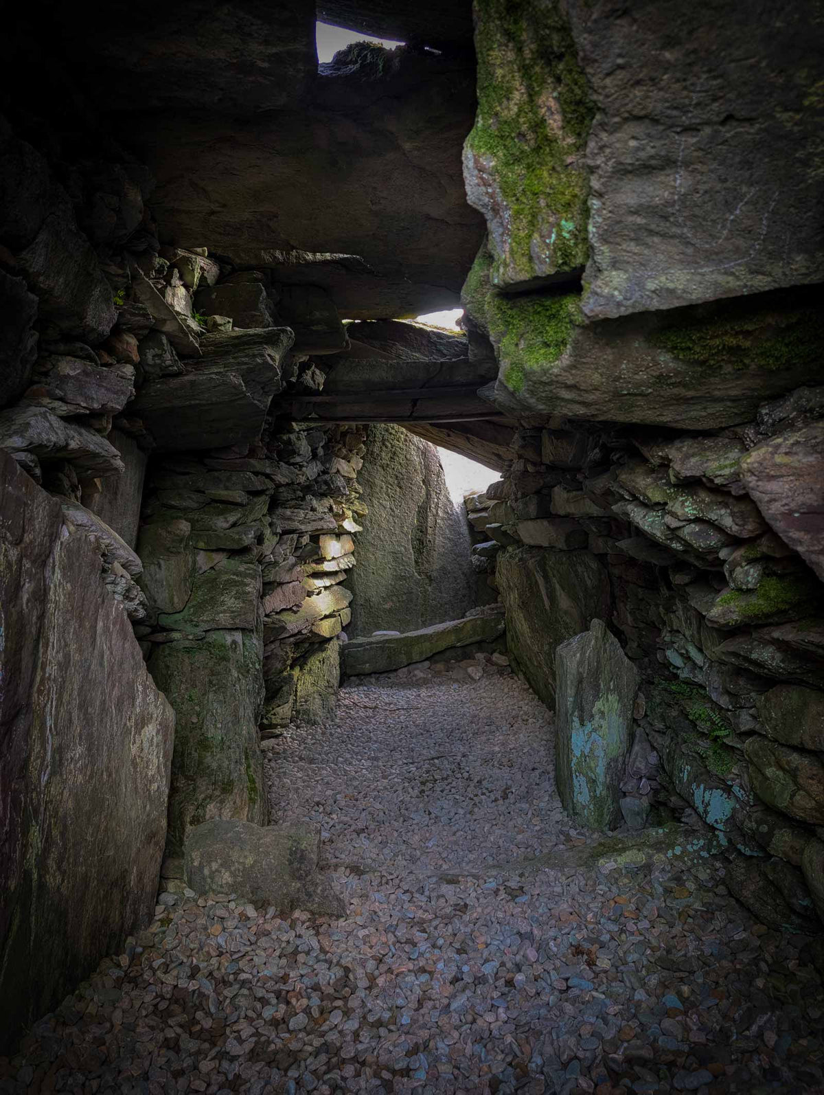 A narrow cairn interior with mossy rock walls and a gravel floor, framed by large boulders under a dark rock ceiling.