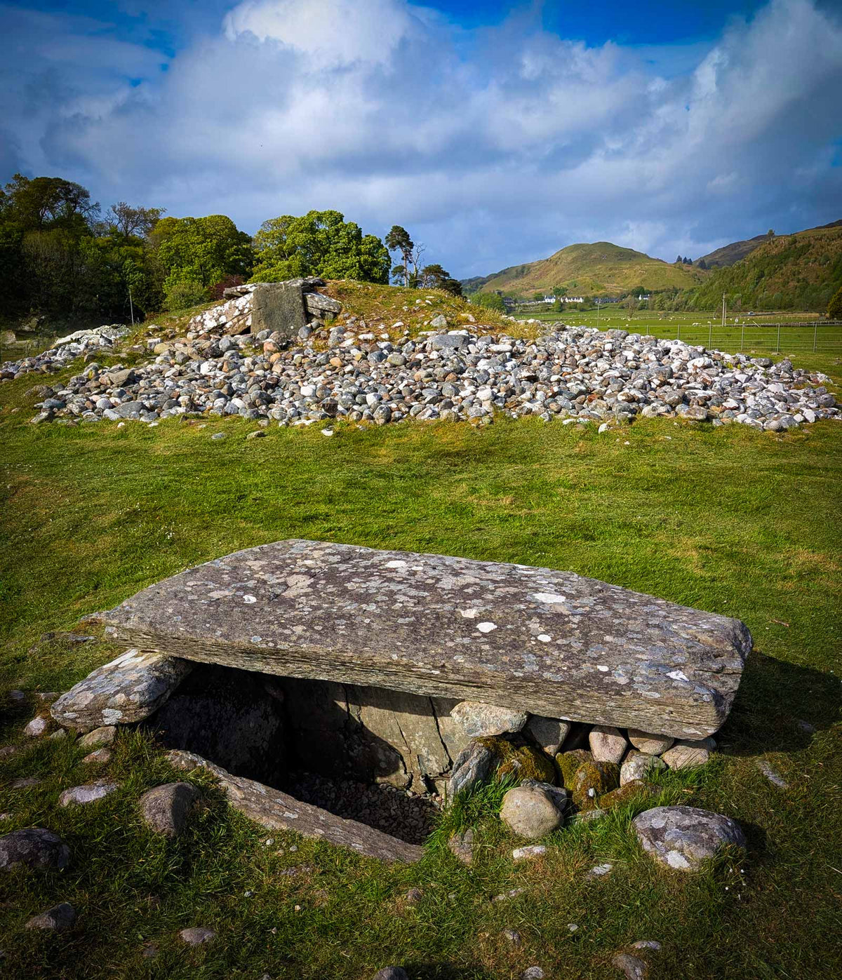 A large stone megalith or ancient tomb structure sits in a grassy field, with more rocks and trees behind it and rolling hills under a blue sky with white clouds.