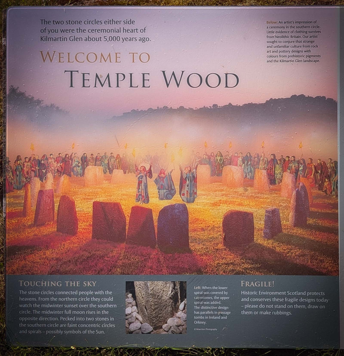 A square printed poster or plaque titled 'Welcome to Temple Wood' showing a sunset scene over stone circles and a crowd of people in ceremonial dress. The design includes descriptive text blocks at the top and bottom and a small inset photo near the lower center.
